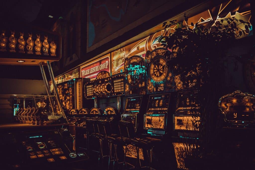 Ambijent kazina, slot aparati u polumraku, ekrani sa brojevima i simbolima, stolice biljka u desnom uglu